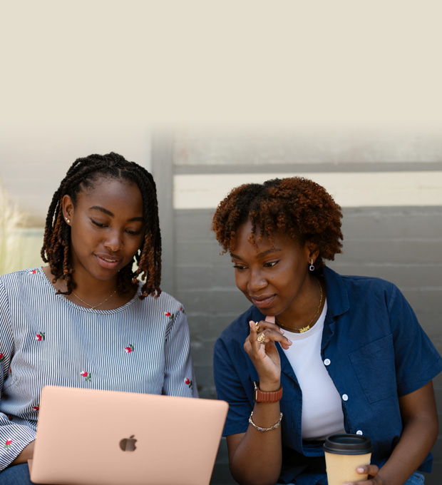 Two women work together on a MacBook on a bench outside.