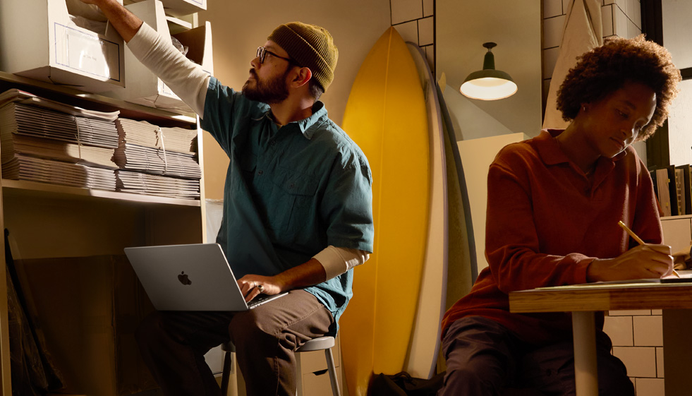 Person in work environment seated on stool using MacBook Air unplugged on their lap while reaching for box on high shelf with other hand