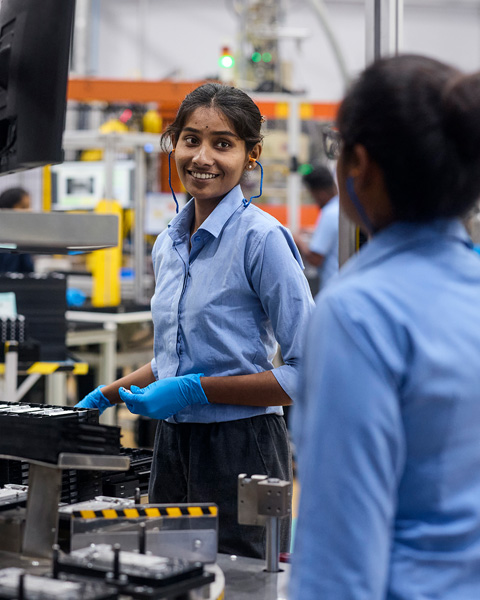 An employee wearing protective earbuds in a component manufacturing facility smiles at a coworker